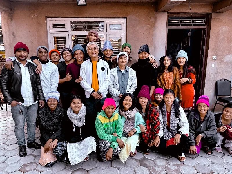 Group of young people standing and sitting outside a building, smiling, wearing colorful winter hats and scarves.