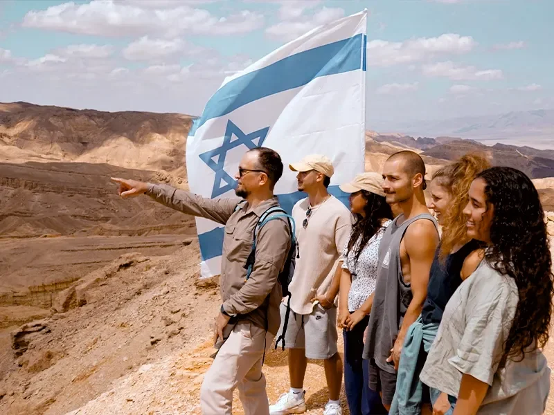 Group of tourists standing in desert landscape with an Israeli flag, one man is pointing into the distance.
