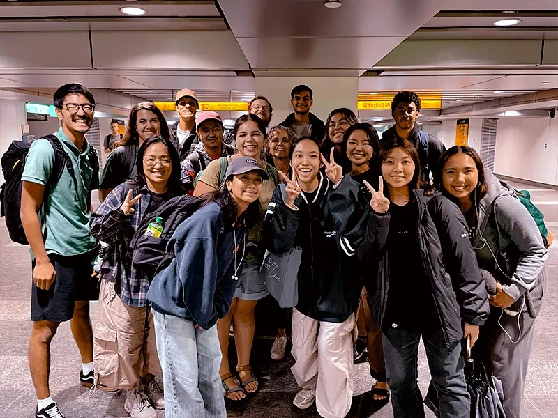 Group of smiling young travelers posing in an airport terminal, some making peace signs.