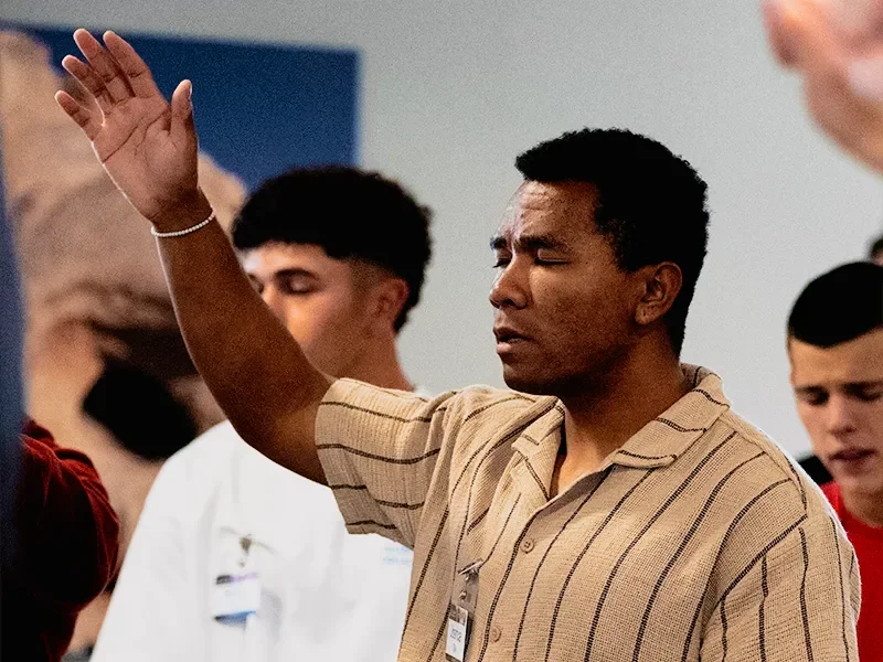 A man with short black hair raising his right hand, eyes closed, possibly praying or in worship, in a room with other young people, some with eyes closed.