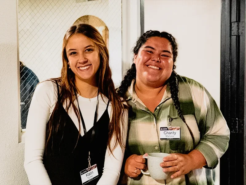Two smiling women, one with long light brown hair and the other with dark curly hair, standing together in an indoor setting. The woman on the right is holding a white mug and wearing a badge that says "Charity" and "Mayette". Both women are wearing badges and casual attire.