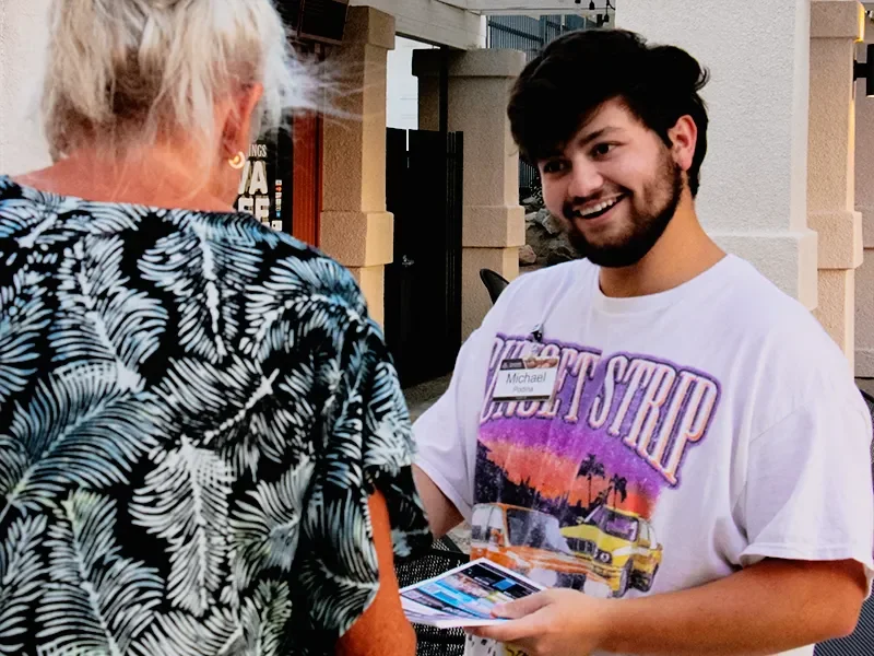 A young man with dark hair and a beard smiling while handing a brochure or flyer to an older woman with blonde hair, wearing a black and white patterned top. They are outdoors with a building in the background.