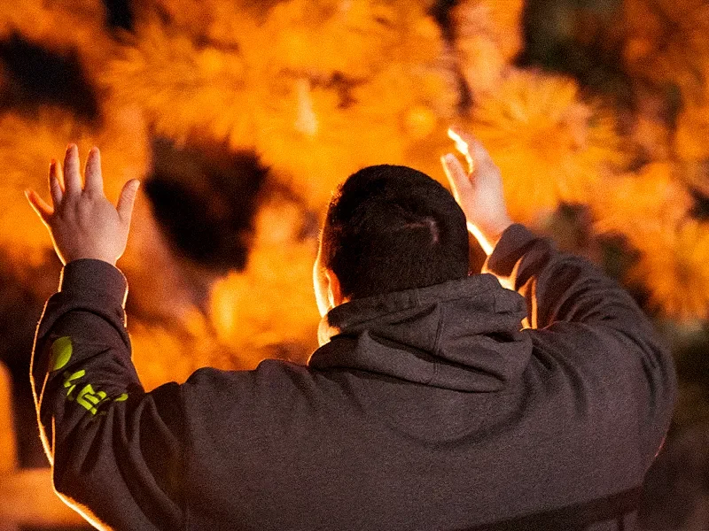 A person with short black hair wearing a dark hoodie is standing outdoors at night, facing away from the camera, with arms raised. Behind them is a large fire with orange flames and dark smoke.
