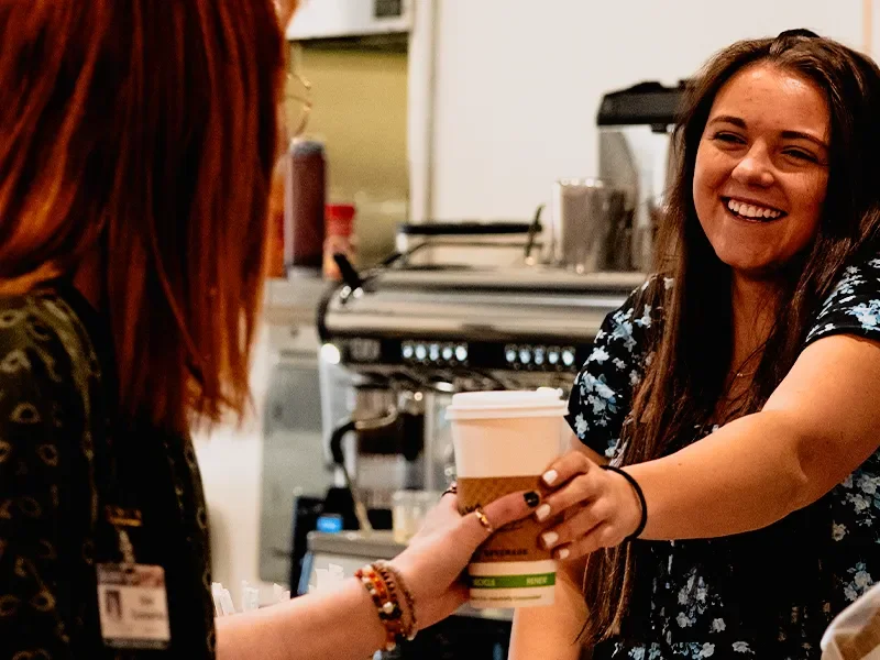 A smiling young woman is handing a coffee to another woman behind the counter in a coffee shop.