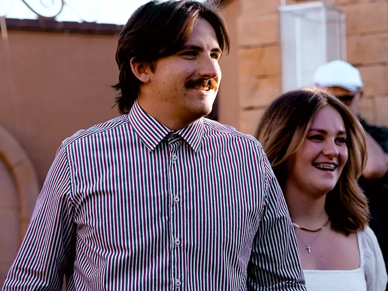 A man with brown hair and a mustache wearing a striped shirt standing next to a smiling woman with brown hair wearing a white top and a necklace, outdoors.