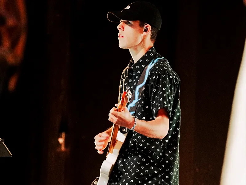 A young man wearing a black baseball cap, black patterned short-sleeve shirt, and a bracelet, playing an electric guitar on stage.