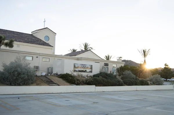 A church with a clock tower at sunset, surrounded by bushes and palm trees, and an empty parking lot in the foreground.