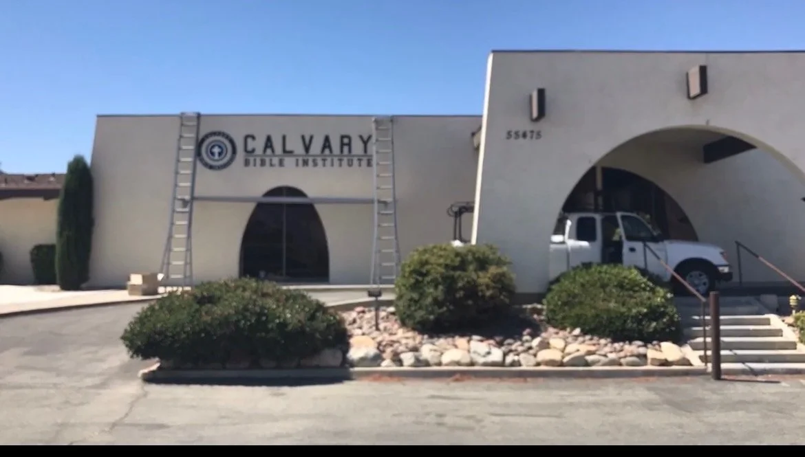 The exterior of Calvary Bible Institute building with a sign, a white pickup truck parked under an arch, bushes, rocks, and a ladder leaning against the building.