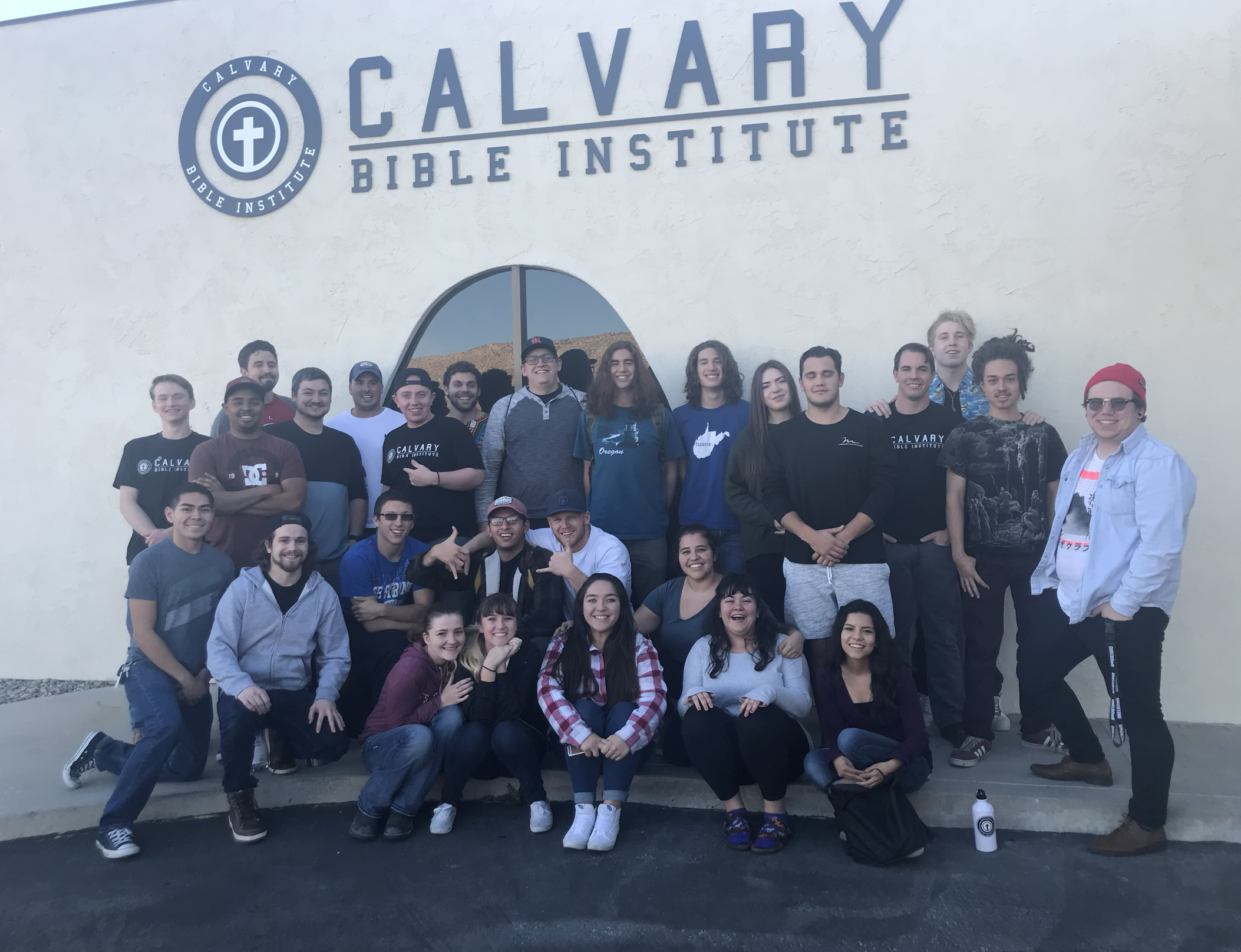 A group of young people standing and sitting in front of a large sign that reads "Calvary Bible Institute" with a logo featuring a cross inside a circle. They are smiling and appear to be part of a school or church group.