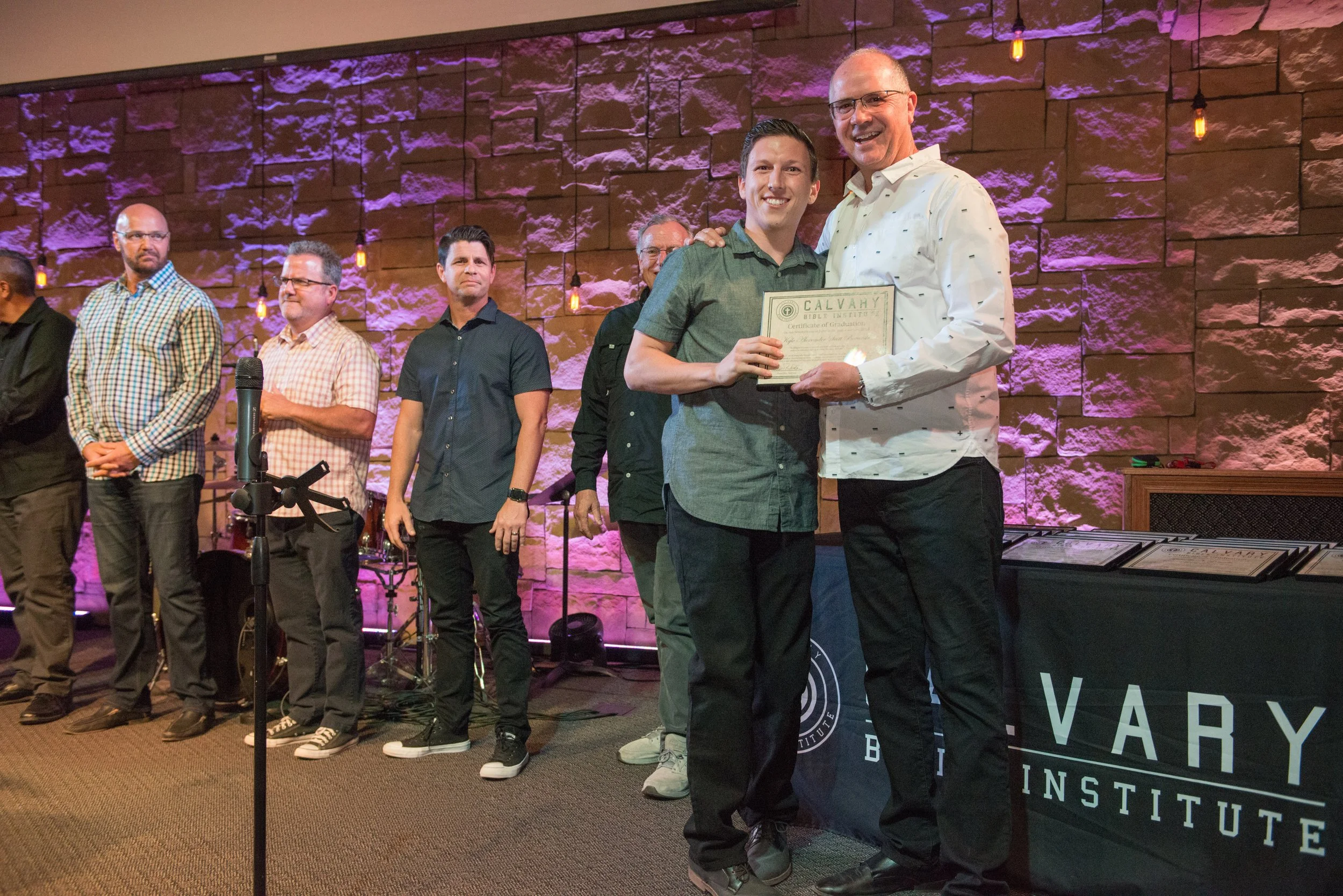 Two men standing together at an award ceremony, one presenting a certificate to the other, with a group of people standing behind them on a stage decorated with purple lighting and a brick wall.