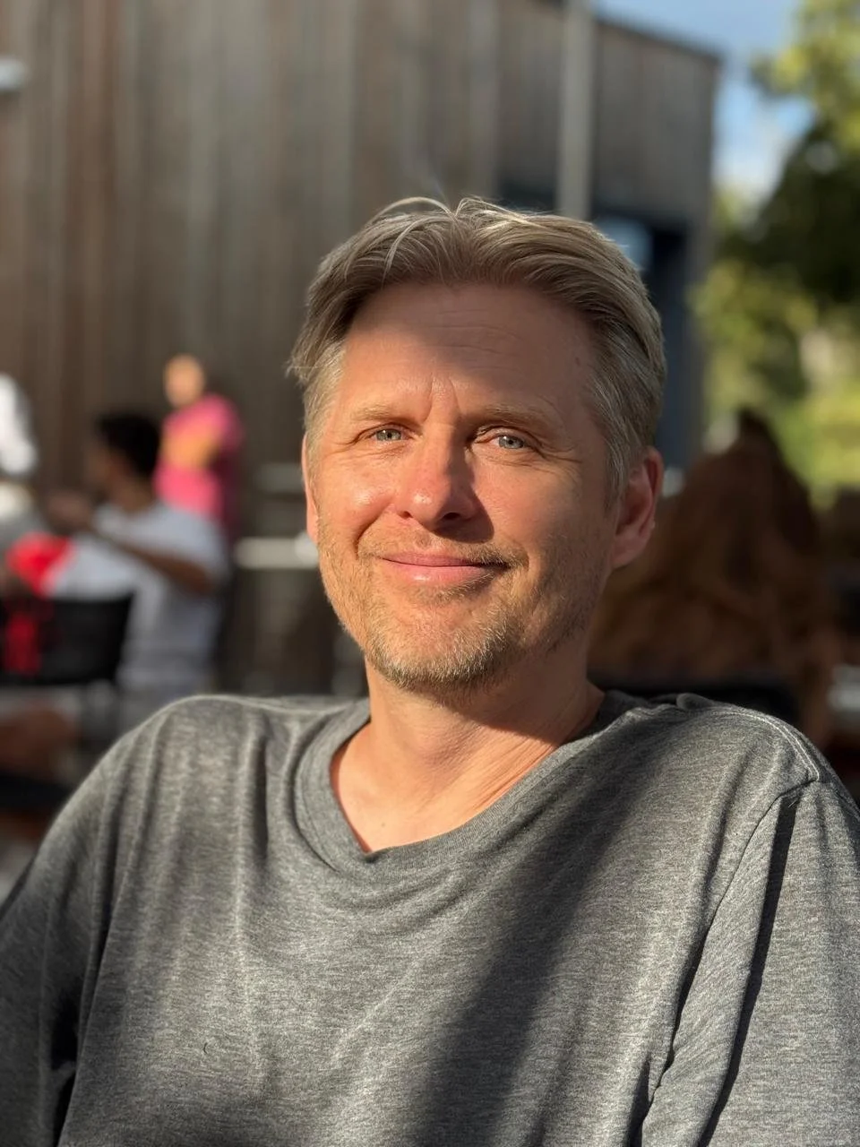 A smiling middle-aged man with blonde hair and a light beard, wearing a gray T-shirt, sitting outdoors during daytime with a blurred background of people and wooden structures.