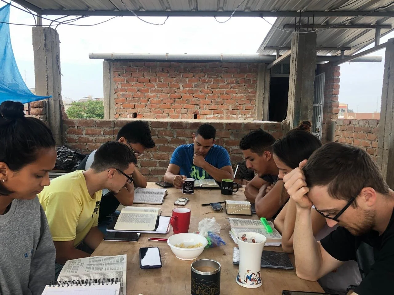 Group of eight young people sitting around a wooden table, reading and studying from books, with cups and snacks on the table, in an outdoor space with brick walls and a metal roof.