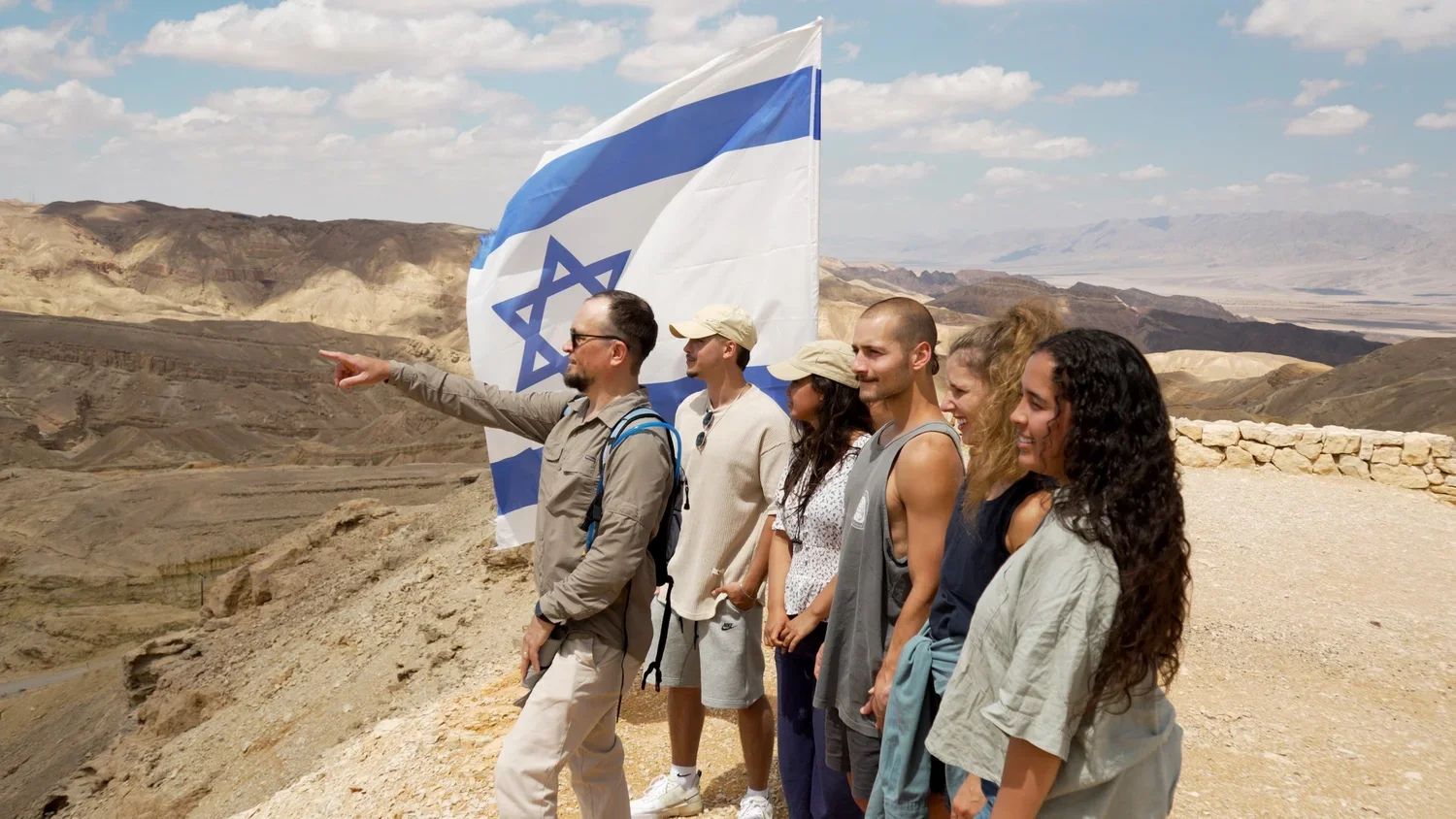 A group of six diverse people standing on a hilltop in a desert with rugged mountains in the background, one person holding an Israeli flag, all smiling and looking towards the same direction.