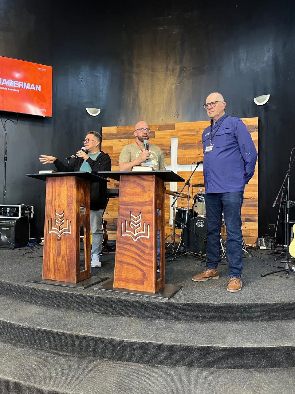 Three men standing on a stage with a black wall background, speaking into microphones, with musical instruments and equipment behind them. One man is wearing glasses, a black jacket, and pointing, another in a beige T-shirt, and the third in a blue shirt and glasses. A large red screen on the left displays the words 'MAGERMAN' and 'Bible Institute'.