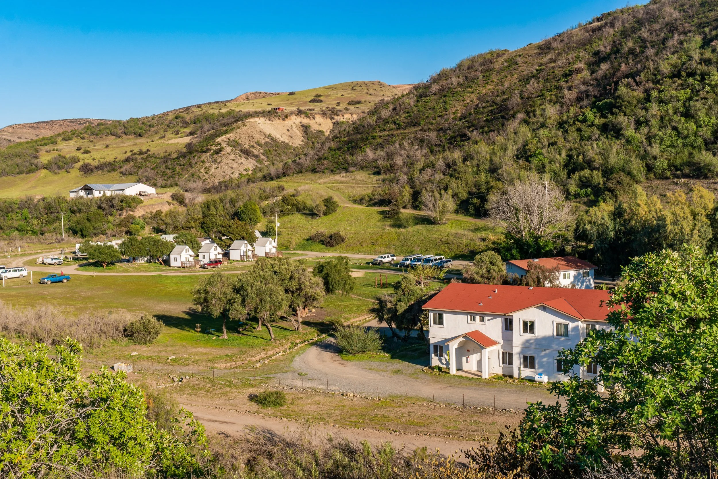 A suburban neighborhood with white houses and red roofs, trees, and parked cars in front of a hillside with sparse vegetation and a clear blue sky.