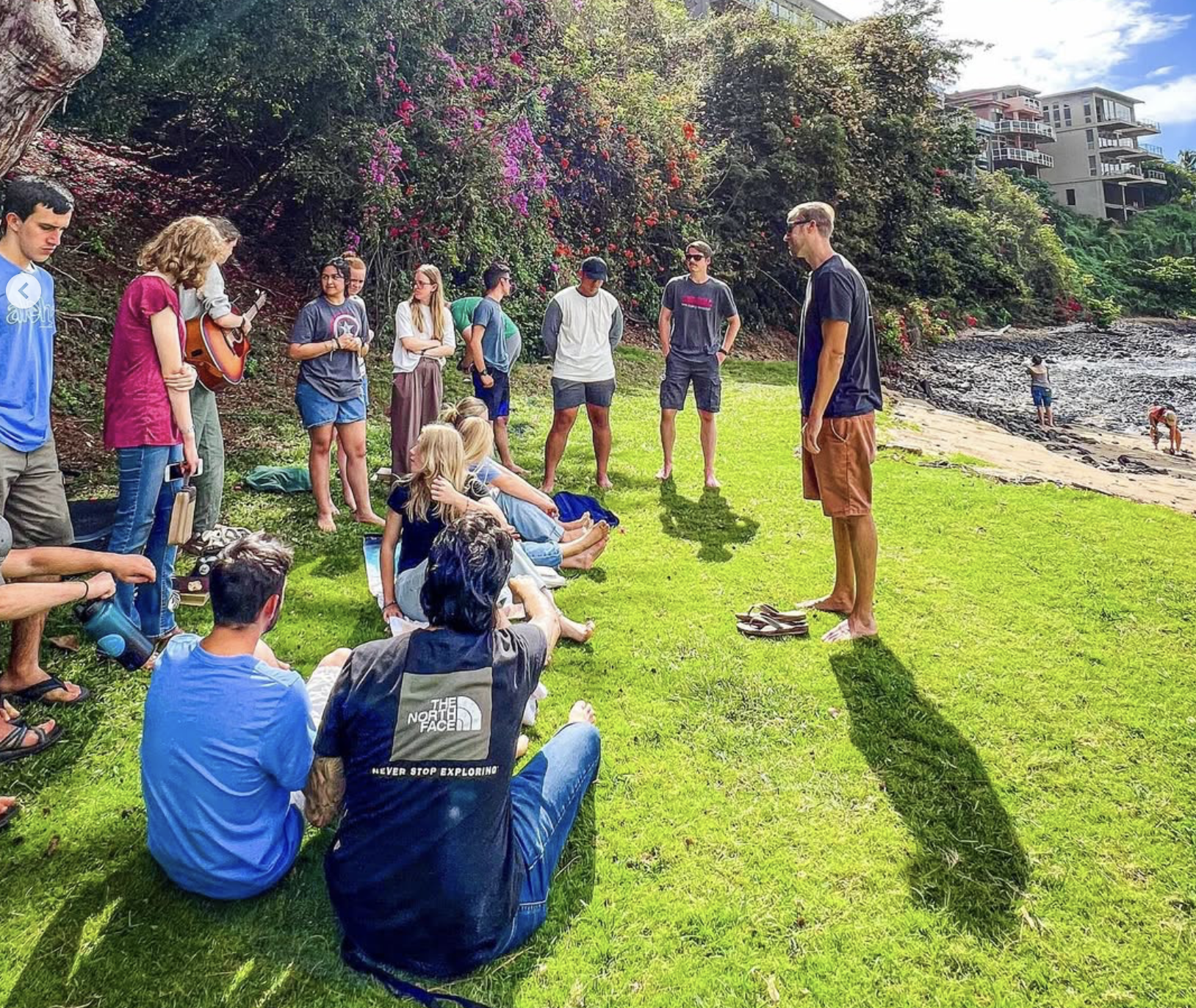 Group of people gathered on a grassy lawn near the beach, some sitting, some standing, listening to a man in black shirt and brown shorts. Others have shoes off and are near rocky shoreline with people swimming in the water. Bright, sunny day with colorful flowers and buildings in the background.