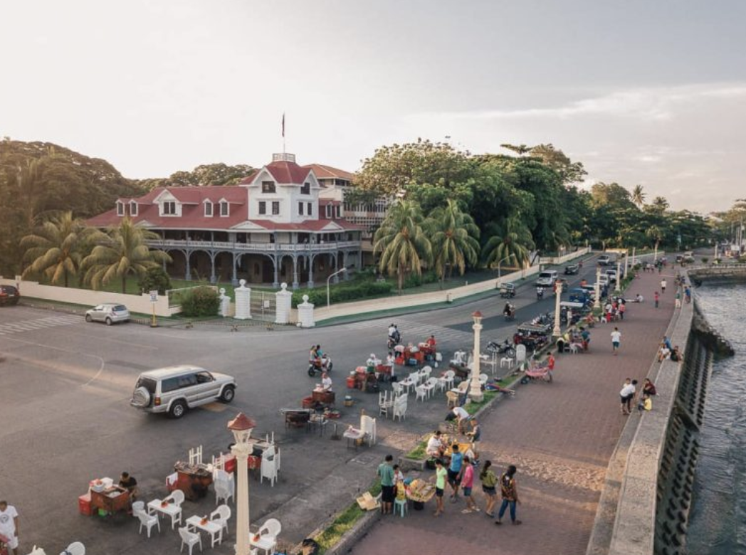 A waterfront promenade with vendors selling goods, people walking, cars driving, a large historic house with a red roof, and palm trees, near a body of water on a partly cloudy day.