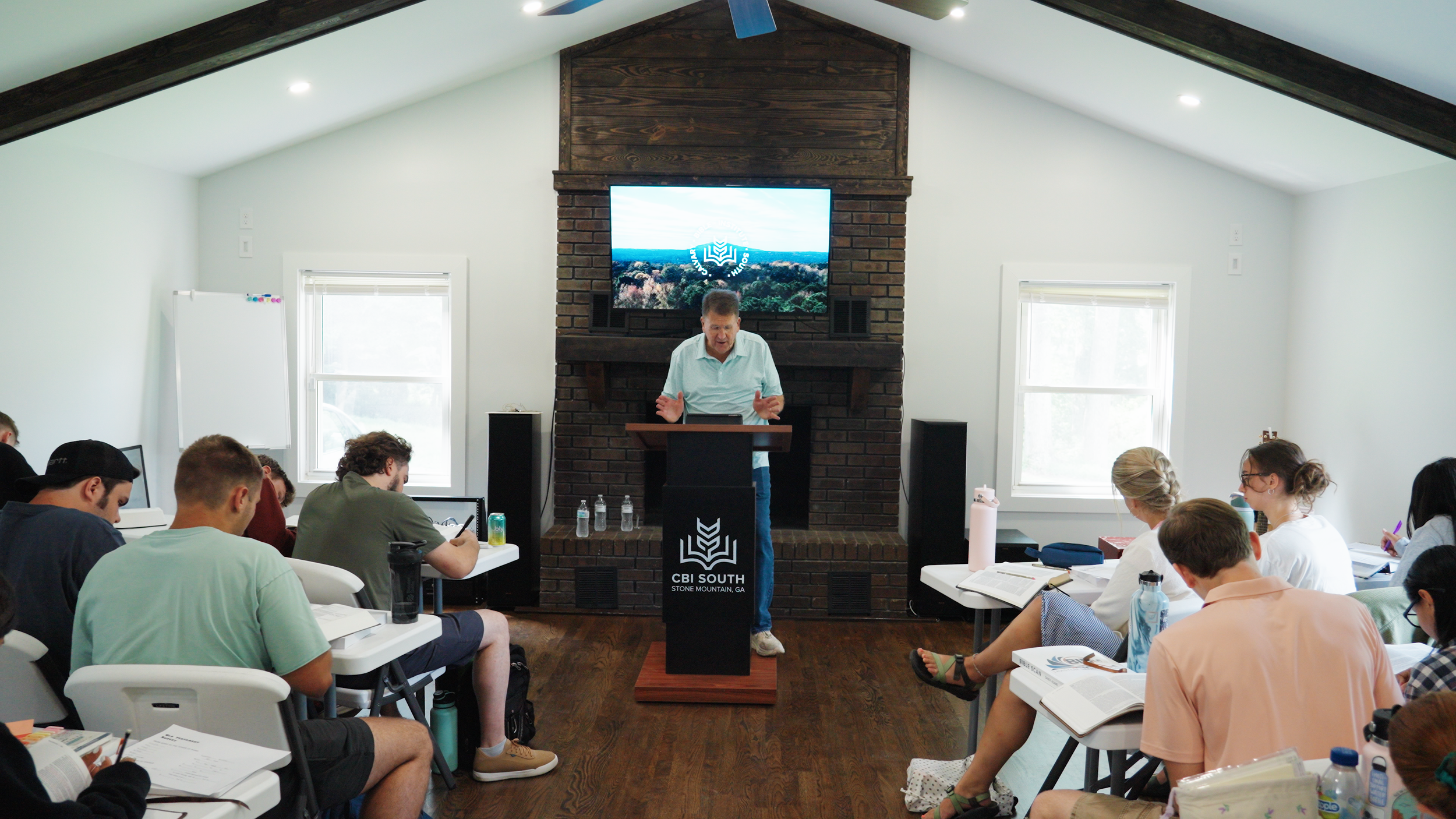 A classroom setting with students seated at white tables, listening to a male instructor at a podium in front of a brick and wood fireplace. A large screen behind him displays the logo of CBI South, Stone Mountain, GA. The room has white walls, two windows, and a sloped ceiling with recessed lighting.