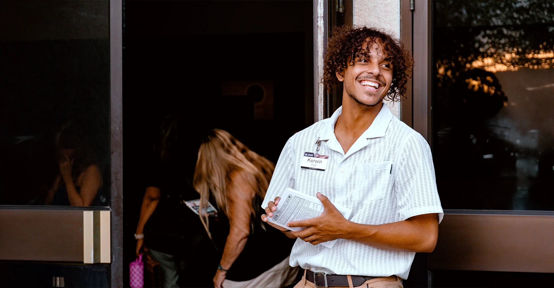Young man with curly hair smiling, wearing a white shirt with a name tag that says 'Kerwin', standing outside a building, holding a brochure, with two women in the background.