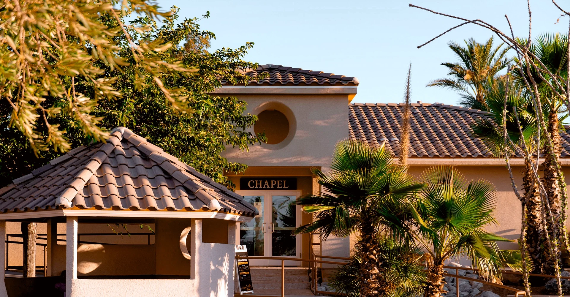 Exterior of a chapel building with a tan stucco facade, a sign that reads "Chapel," and a tiled roof. There are trees and palm plants around the building, with a small pavilion in the foreground.