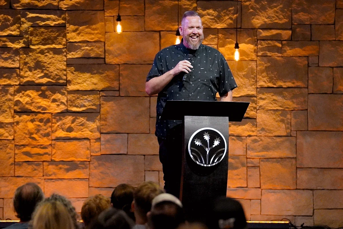 A man with a beard, wearing a black shirt with small white patterns, smiling and holding a microphone while standing at a podium. The background is a stone wall with warm lighting and hanging light bulbs. An audience is partially visible in the foreground.