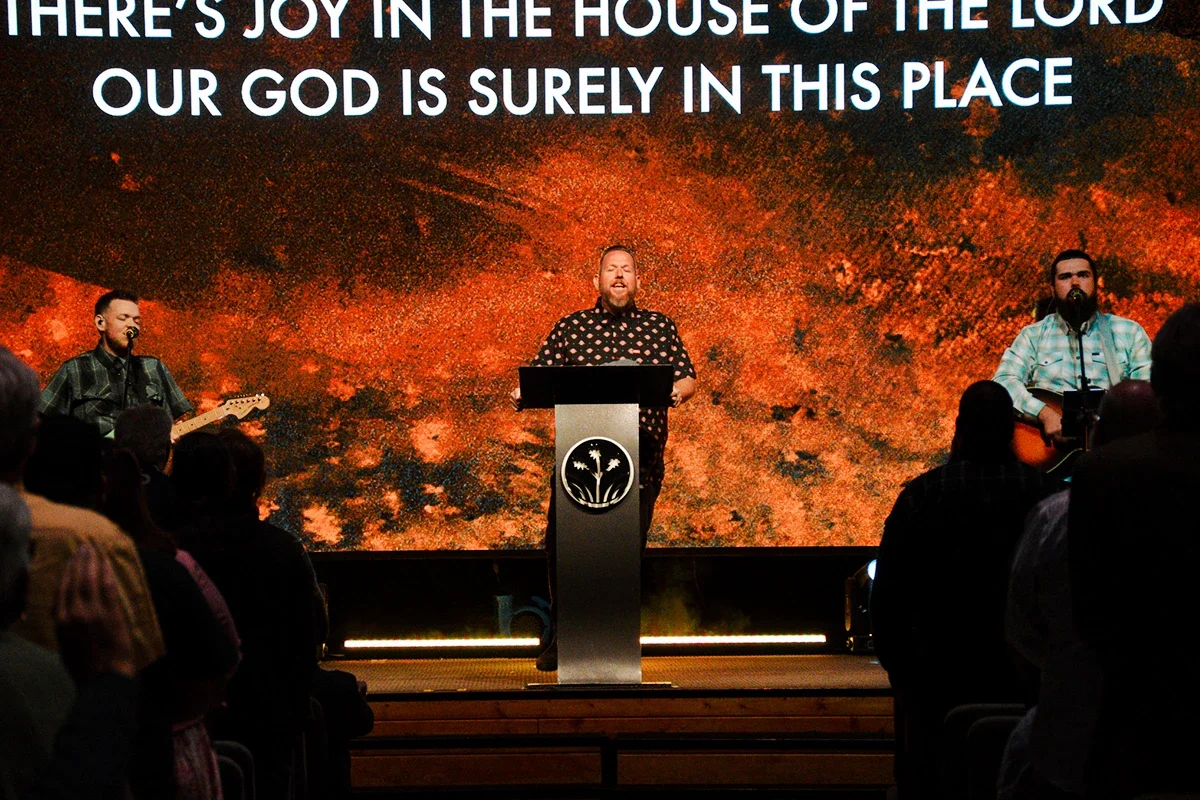A man is speaking at a podium with a logo, with two musicians playing guitar on either side, in front of a large screen displaying the words 'THERE'S JOY IN THE HOUSE OF THE LORD OUR GOD IS SURELY IN THIS PLACE'. Audience members are visible, facing the stage.
