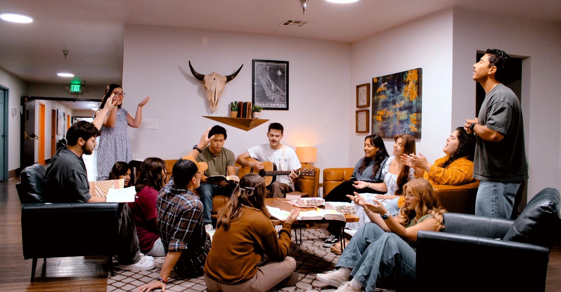 A group of young people gathered in a cozy living room, some sitting on the floor, others on couches, engaged in singing, playing guitar, and worshiping together. Some individuals have their hands raised or are clapping, creating an atmosphere of worship and fellowship. The room has modern decor with artwork, a mounted animal skull, and a bookshelf on the wall.