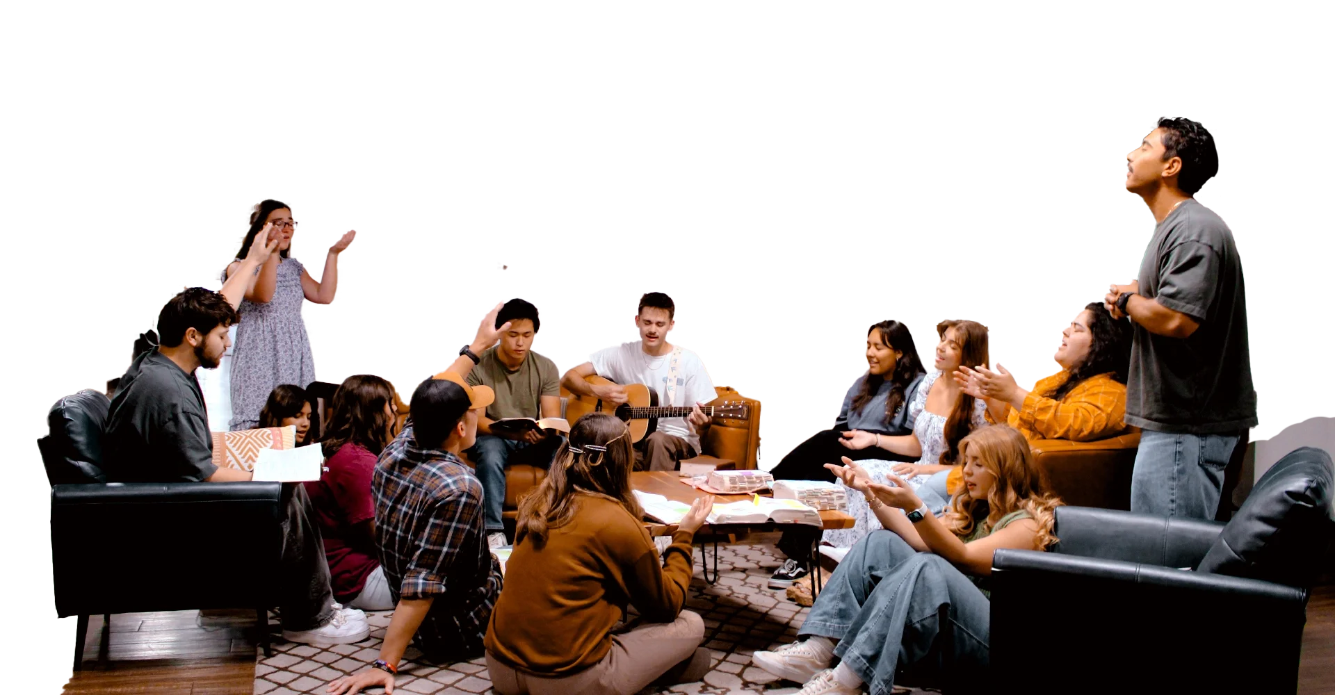A group of people gathered in a living room, some sitting on sofas and chairs, participating in a discussion or music session, with one person playing guitar, and others engaging in singing and clapping.