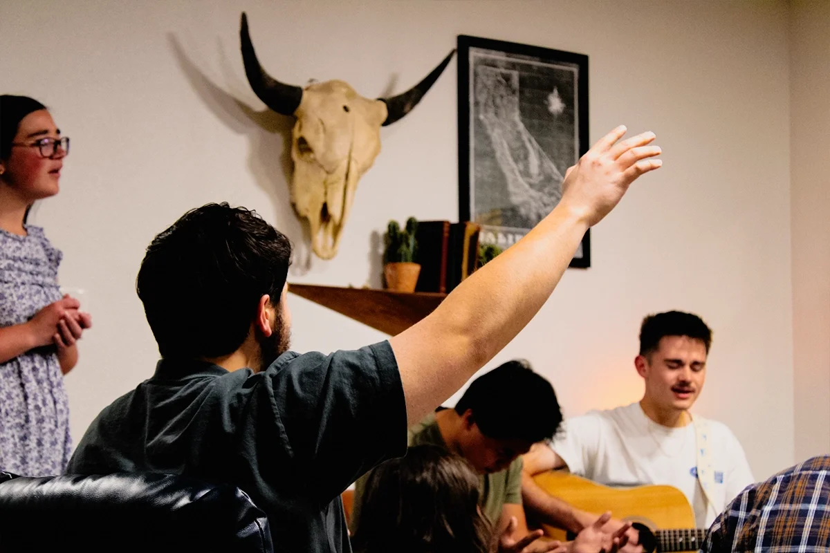 People gathered in a room, with one person raising his hand. A woman stands to the side. There is a buffalo skull mounted on the wall, along with a framed picture and a shelf with cacti and books.