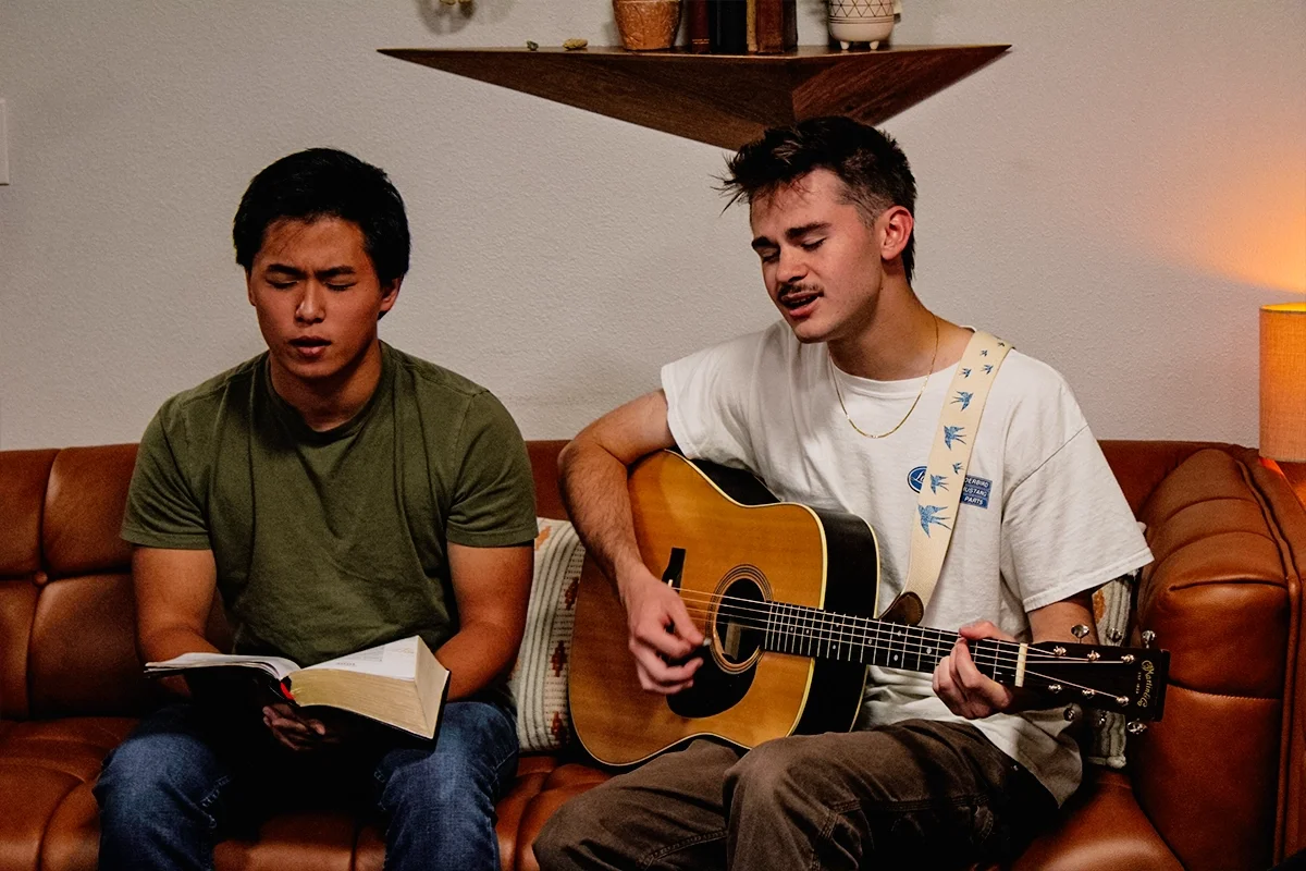 Two young men sitting on a brown leather couch in a cozy living room, one reading a book and the other playing an acoustic guitar.