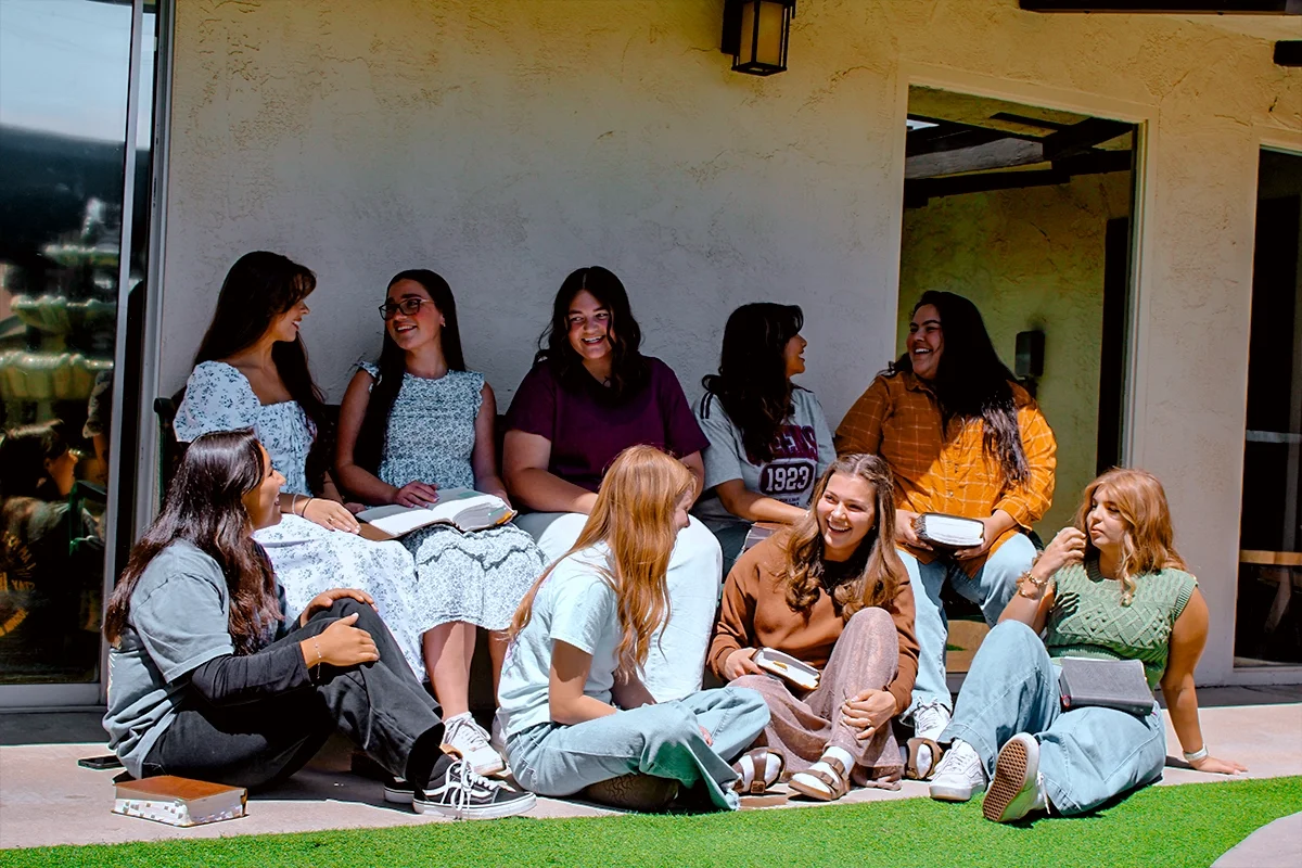 Group of young women sitting and standing outside, engaged in conversation and smiling, some holding books.