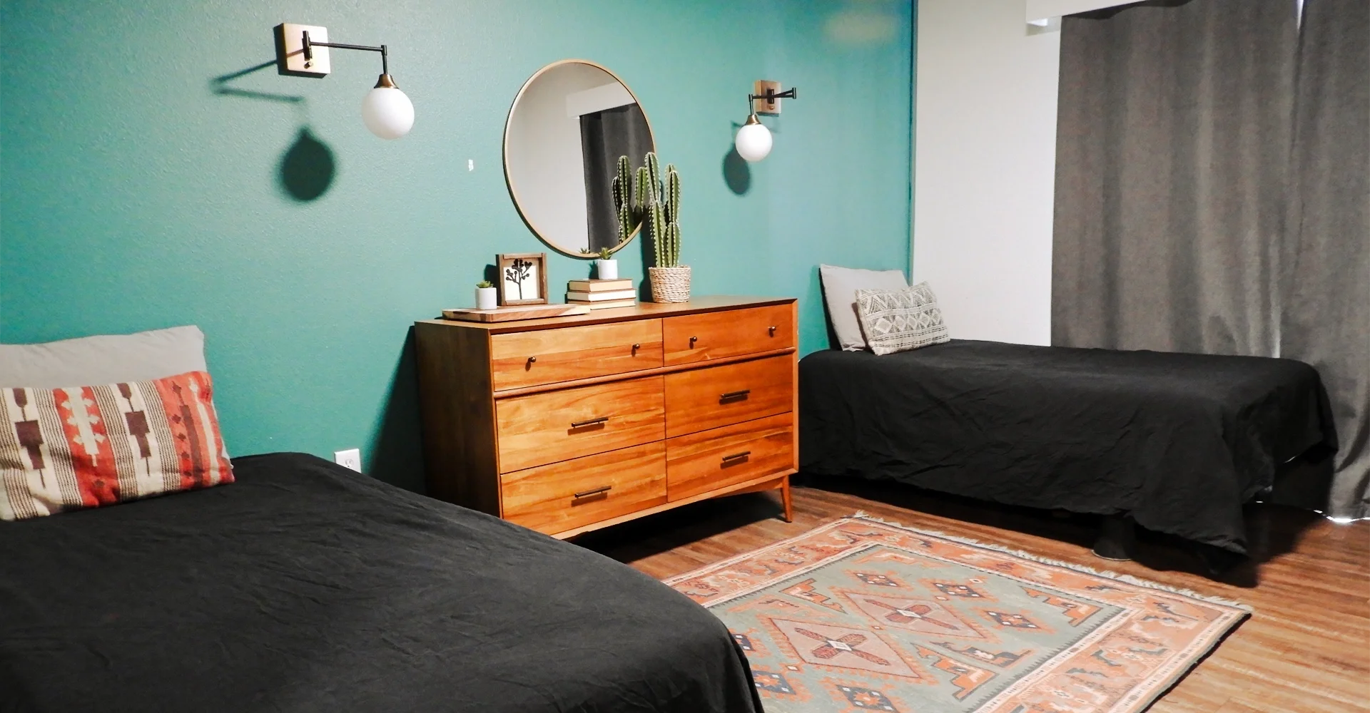 Bedroom with teal accent wall, two beds with black bedding, a wooden dresser with plants and books, an oval mirror, and wall-mounted lights.