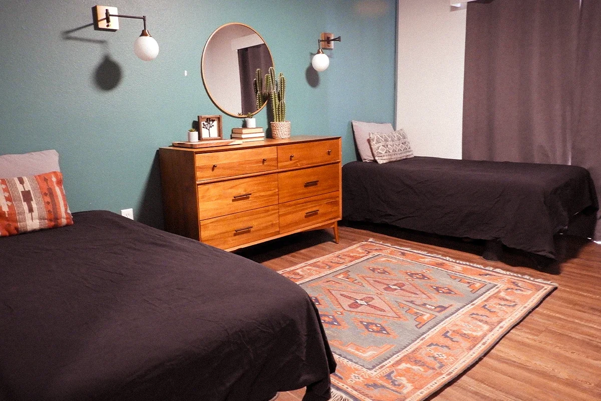 A bedroom with two black beds, a wooden dresser with a round mirror and a cactus, and a patterned area rug on hardwood floor.