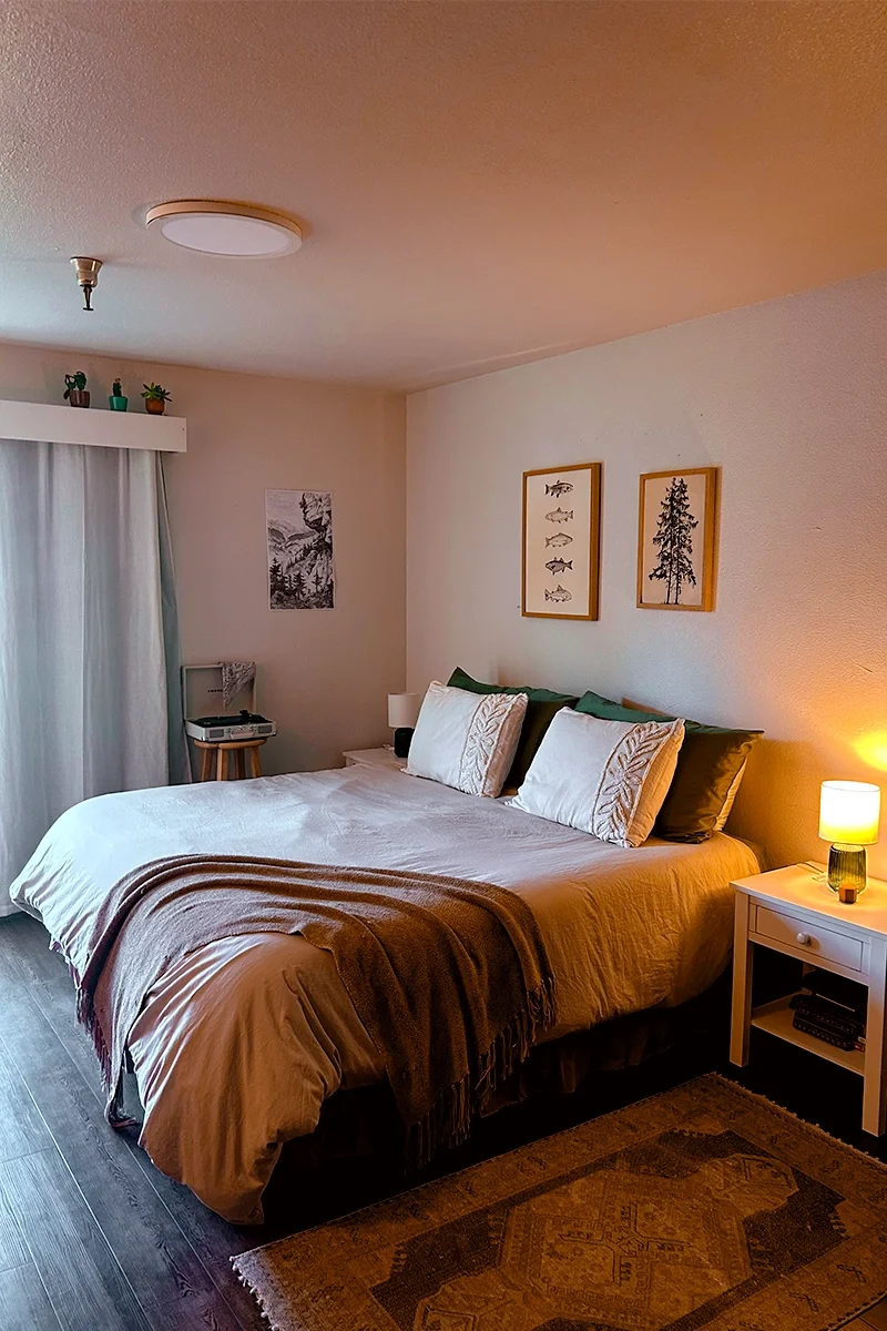 A bedroom with a bed featuring white and dark green pillows, a beige blanket, and a brown throw. There is a nightstand with a lamp and a few books, and a window with white curtains. Decorations include framed pictures of fish and trees, and small potted plants on a shelf near the ceiling.