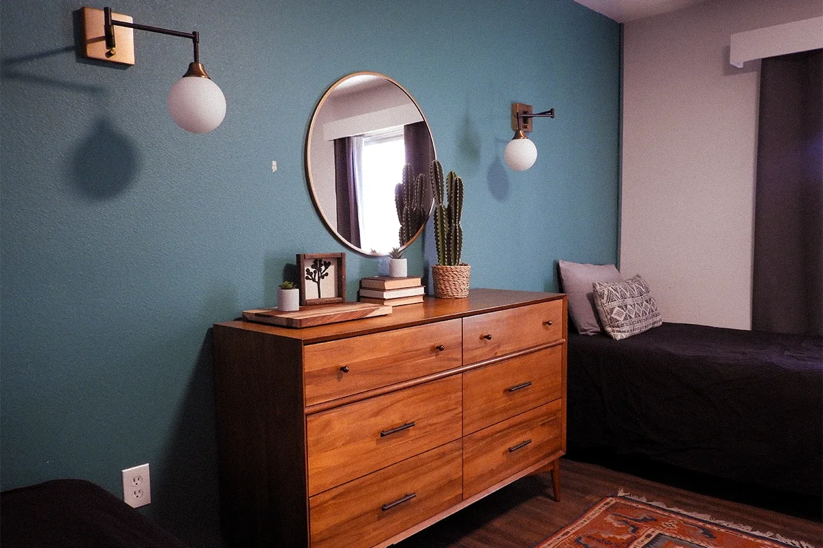 A bedroom interior with a teal accent wall, a round mirror, and wall-mounted light fixtures. A wooden dresser with decorative items such as potted plants, framed art, and a stack of books is in front of the wall. A bed with dark bedding and patterned pillows is on the right. A window with curtains is reflected in the mirror, and part of a colorful rug is visible at the bottom of the image.