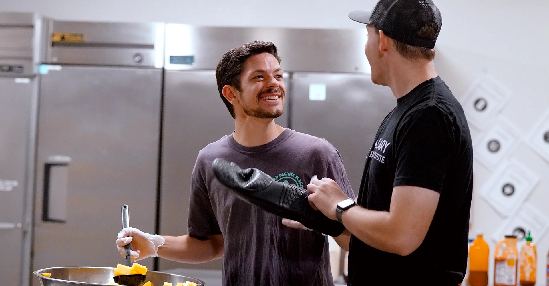 Two men in a kitchen, one smiling at the other. The man on the left is holding a spatula in a bowl with chopped vegetables, wearing gloves. The man on the right is wearing a black t-shirt, a cap, and a smartwatch, talking to the other.