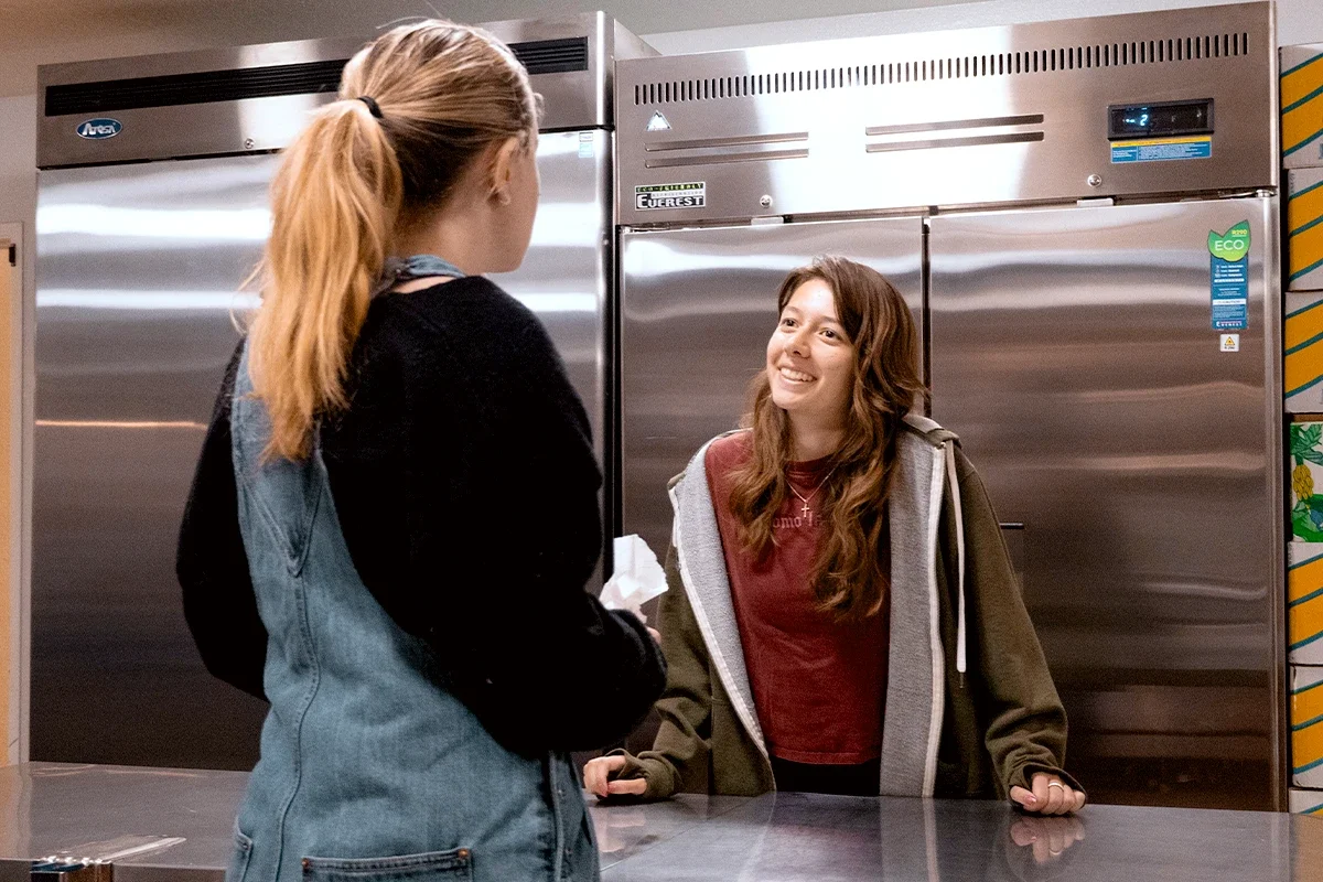 Two young women are having a conversation in a commercial kitchen or store. One woman is standing with her back to the camera, wearing a black and denim jacket, while the other woman is facing her, smiling. The woman facing the camera has long brown hair, wears a red shirt and an olive green zip-up hoodie.