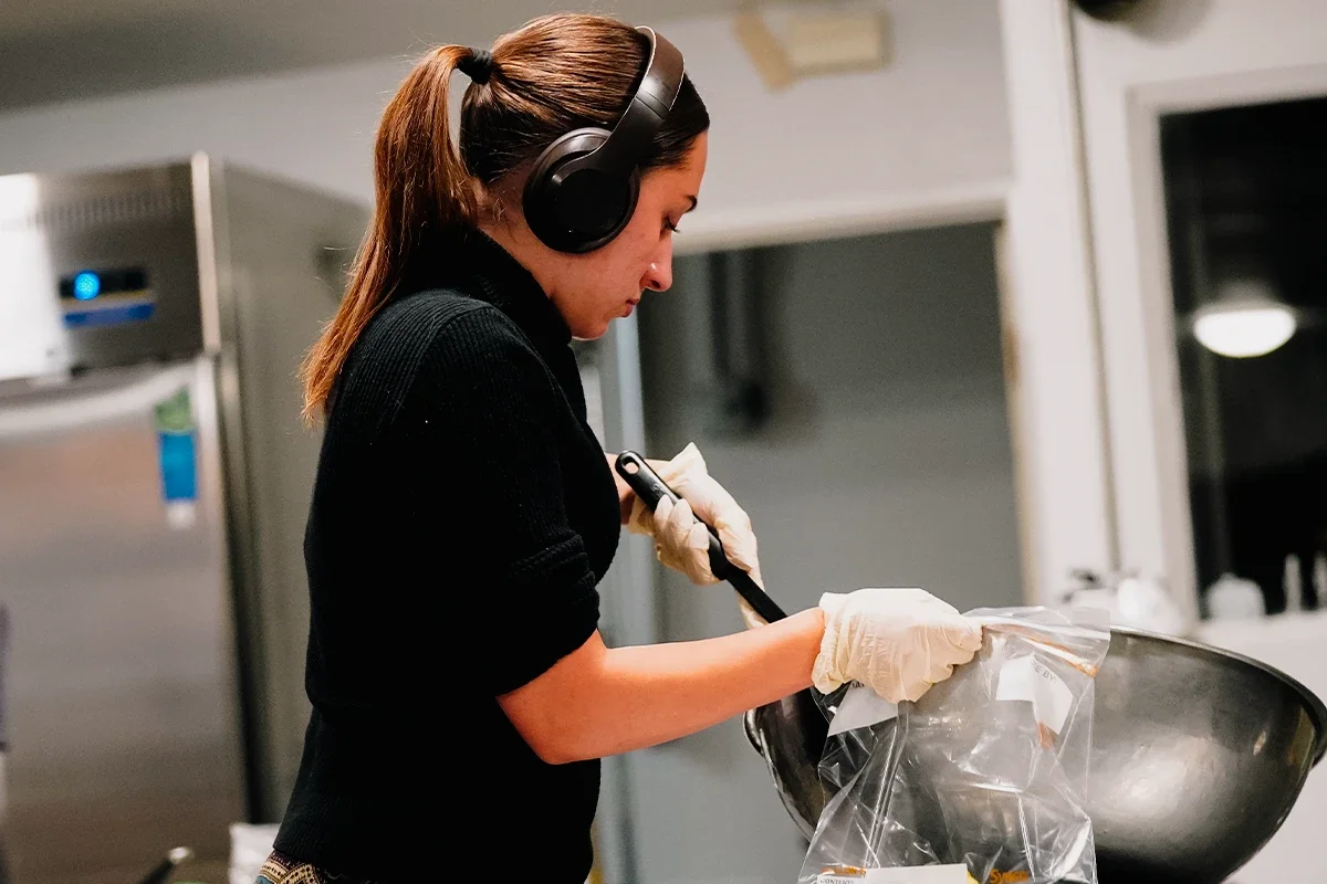 A woman wearing headphones and gloves, working with a metal mixing bowl and a spatula in a kitchen.