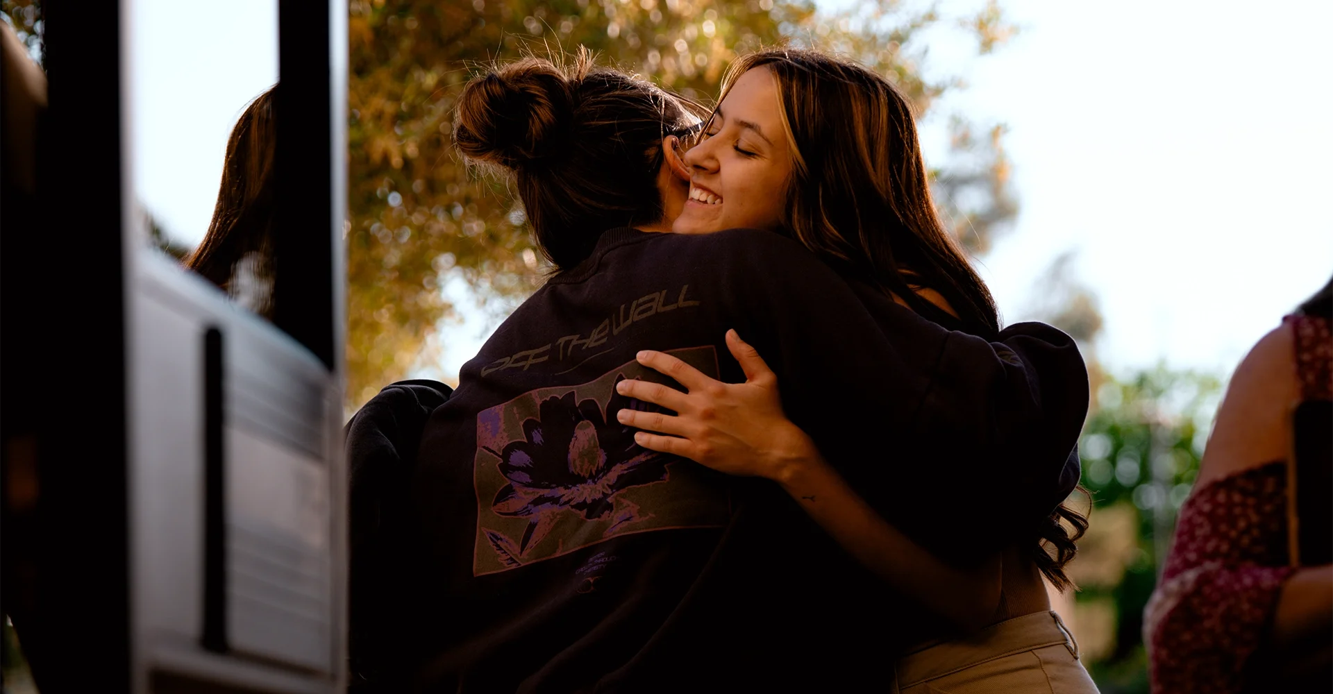 Two women hugging outdoors during daytime, smiling with eyes closed, with trees and sunlight in the background.