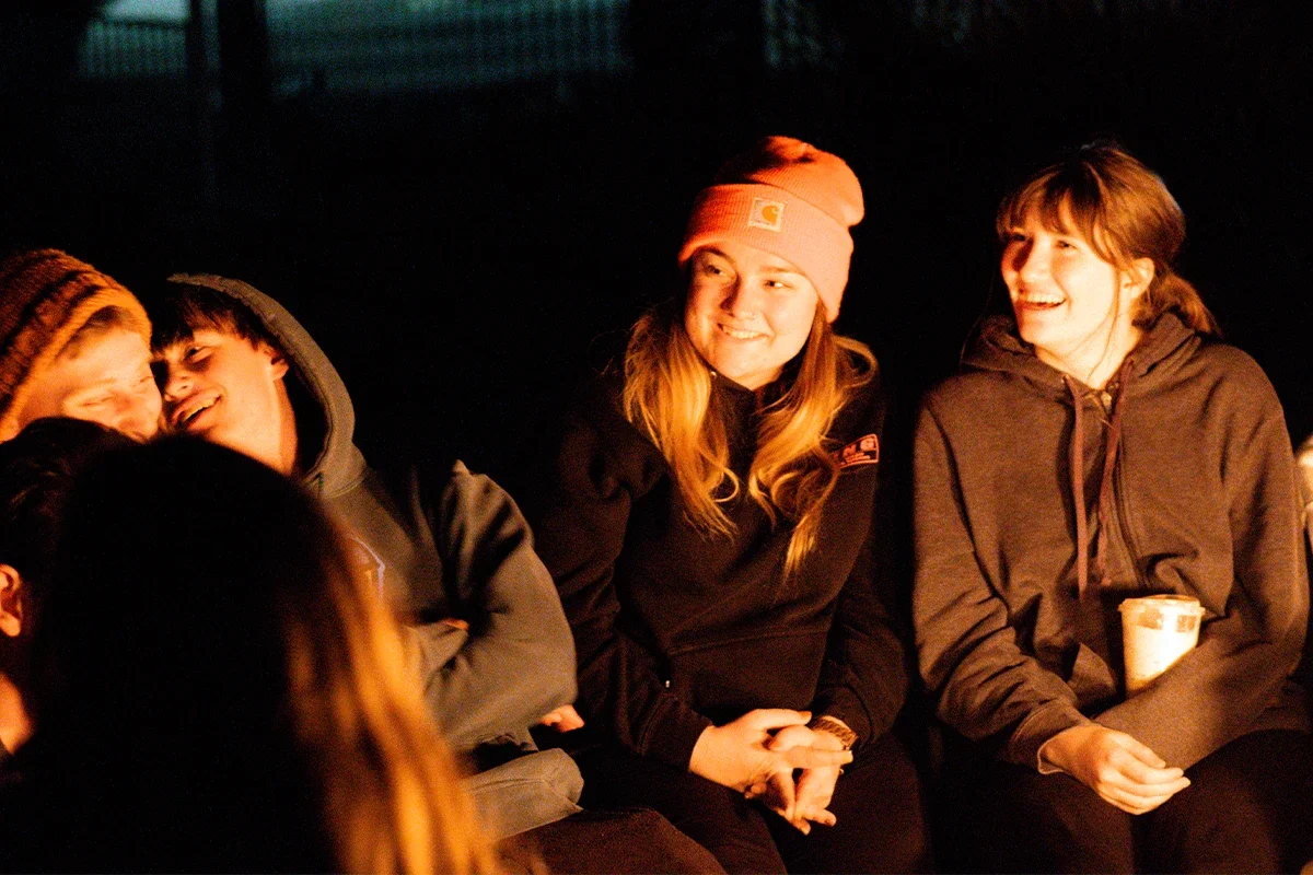 A group of four young people sitting outdoors at night, smiling and laughing around a campfire.