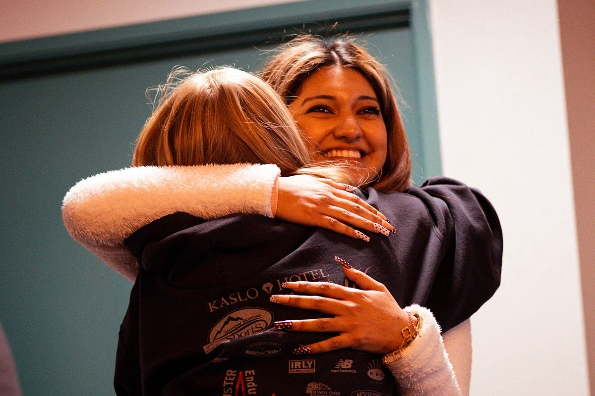 Two women hugging each other warmly, smiling, one with brown hair and the other with reddish hair, in an indoor setting.