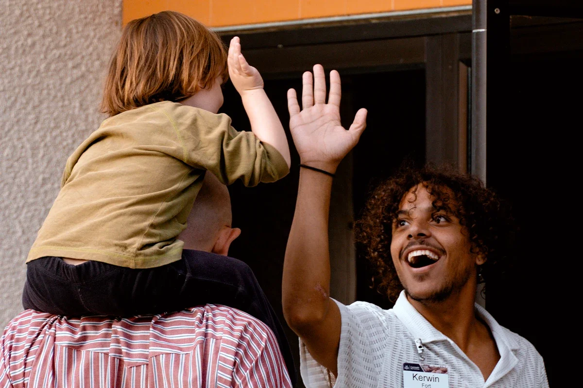 A man with curly hair and a big smile high-fives a young child sitting on his shoulders as they celebrate together.
