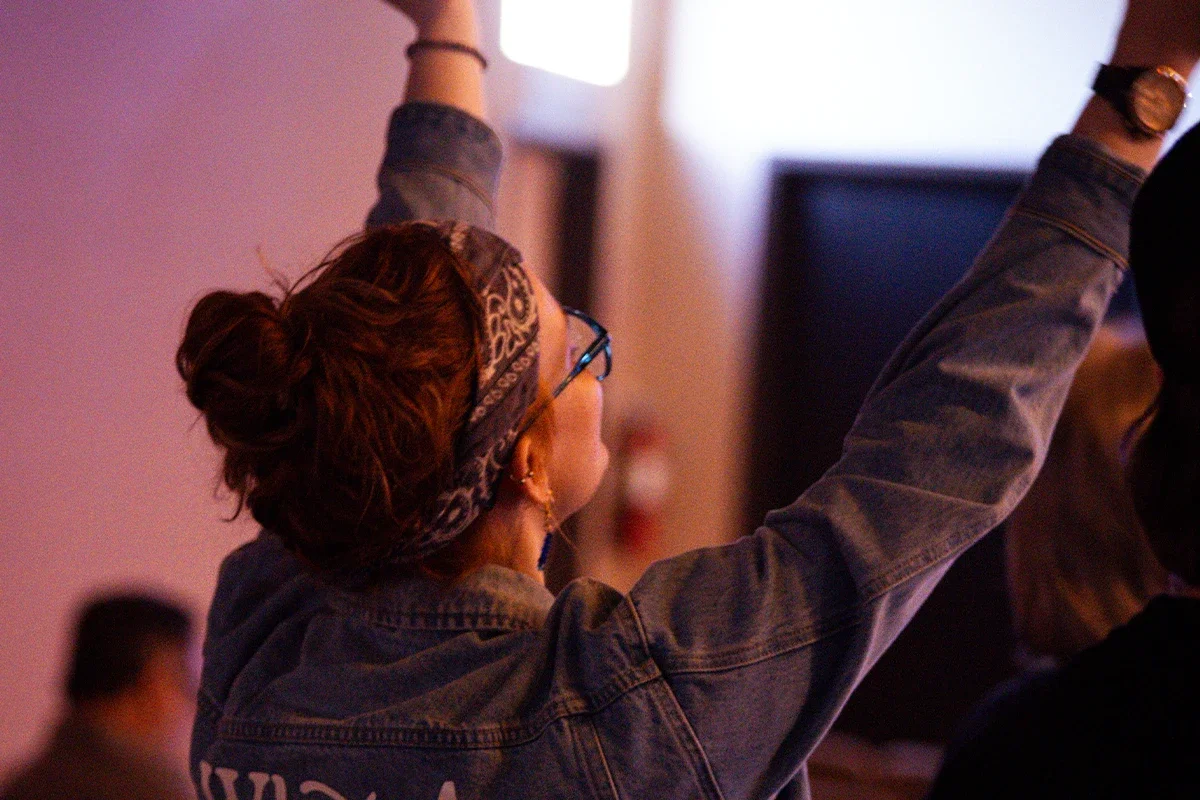 A woman with glasses and earrings wearing a denim jacket and headscarf, raising her arms in an indoor setting.