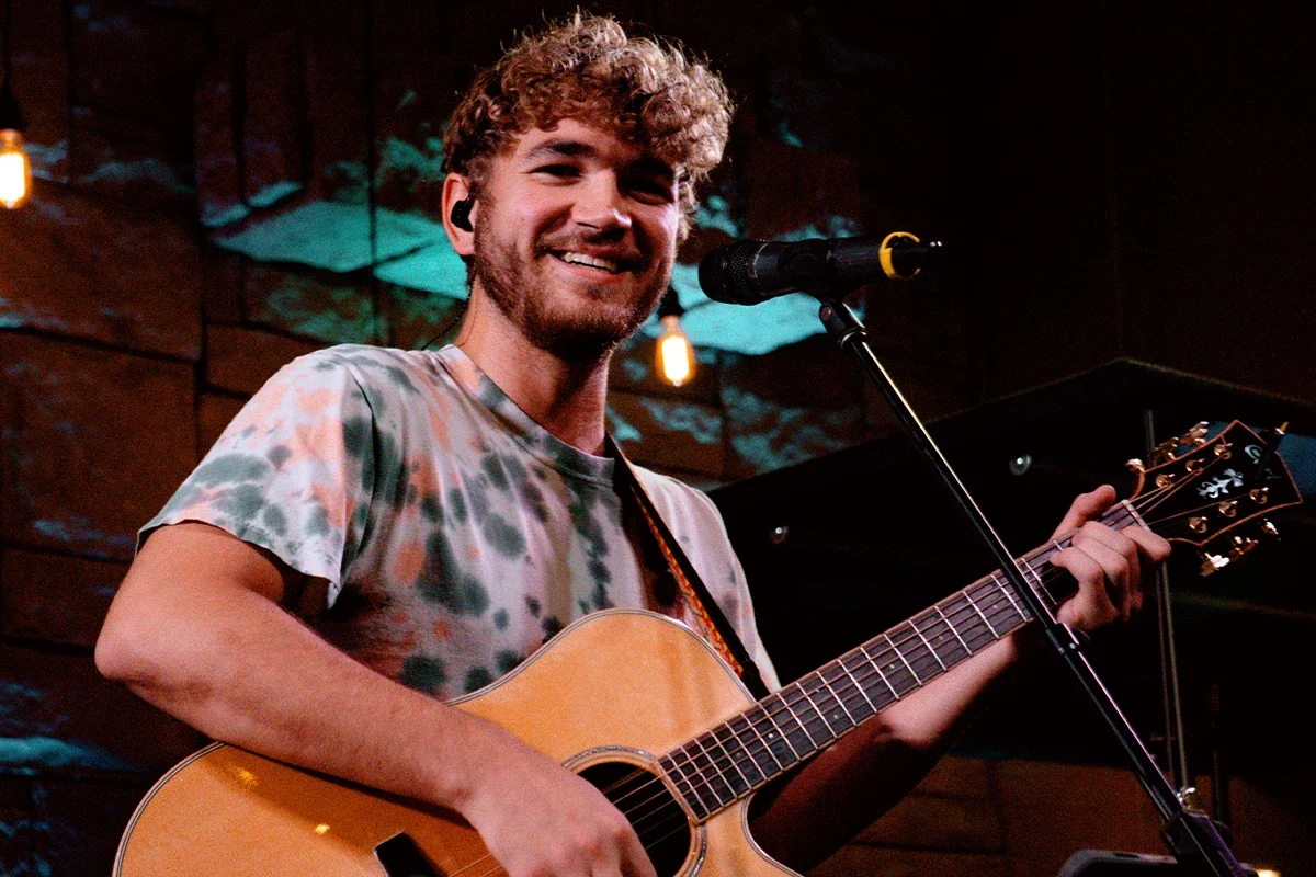 Young man with curly hair smiling, playing an acoustic guitar, performing on stage with a microphone in front of him.