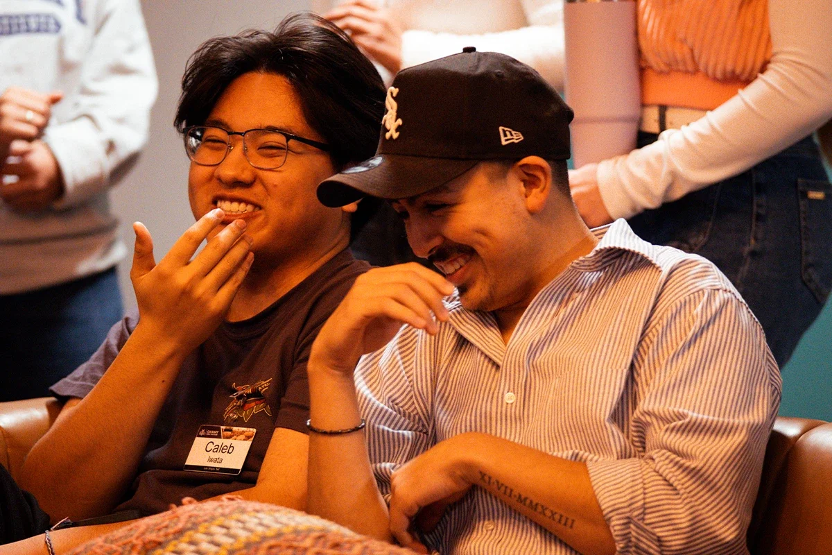 Two young men sitting and laughing together, one wearing glasses and a name tag, the other wearing a black baseball cap and striped shirt.
