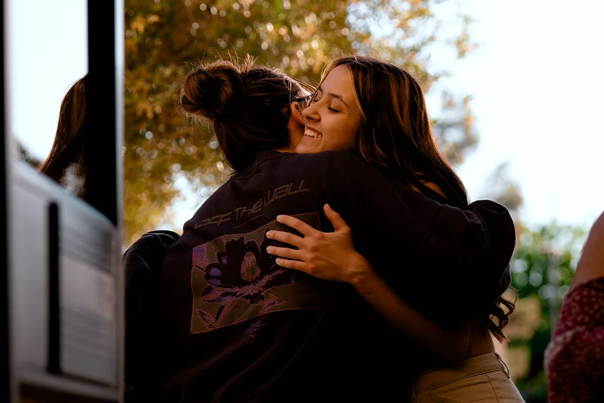 Two women are hugging outdoors, smiling with eyes closed, surrounded by trees and sunlight.