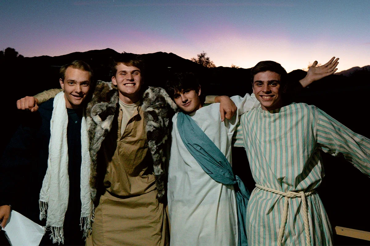 Four young men dressed in biblical costumes standing outdoors during sunset or dusk, with mountains in the background.