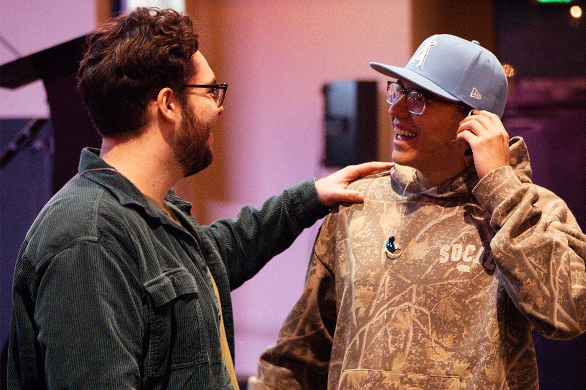 Two men are smiling and talking in a room, one has his hand on the other's shoulder; the man on the left has dark hair, glasses, and a beard, wearing a dark jacket. The man on the right is wearing glasses, a gray cap, and a camouflage hoodie, with an earbud in his ear.