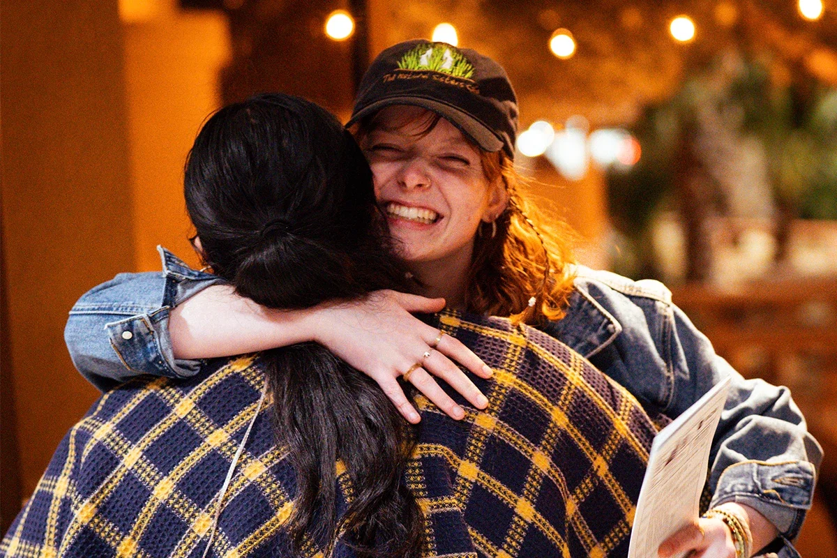 Two women hug, one smiling widely, in a warm, softly lit setting.