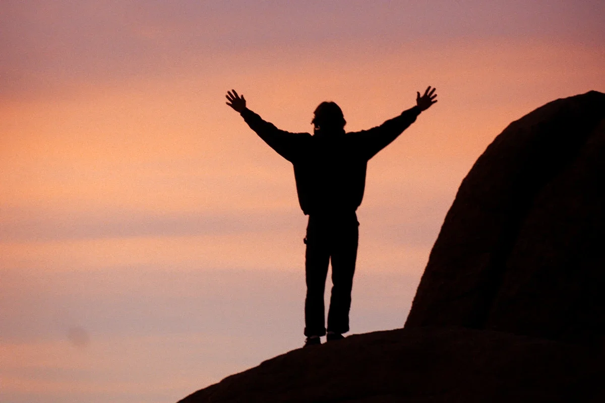 Silhouette of a person with outstretched arms standing on a rocky hill at sunset with a pink and orange sky.